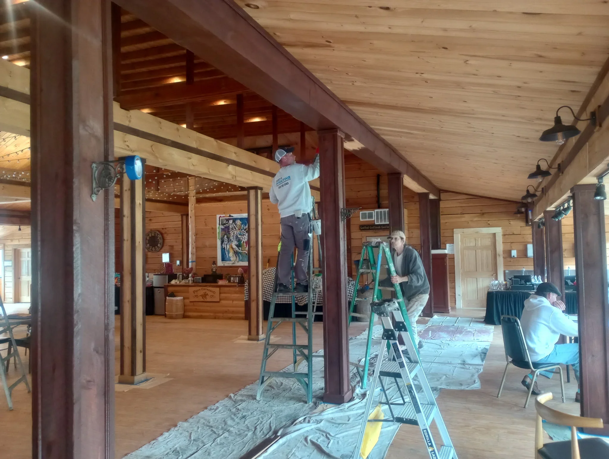 Painters on ladders applying finish to wood ceiling in spacious lodge interior with exposed beams